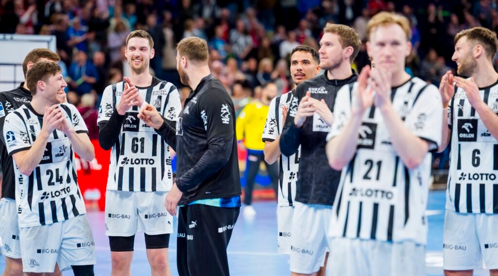 Eine Gruppe männlicher Handballspieler in schwarz-weißen Trikots steht auf einem Spielfeld, klatscht und lächelt, während im Hintergrund eine Menschenmenge zu sehen ist. Einige Spieler sind einander zugewandt und scheinen zu feiern oder etwas anzuerkennen.