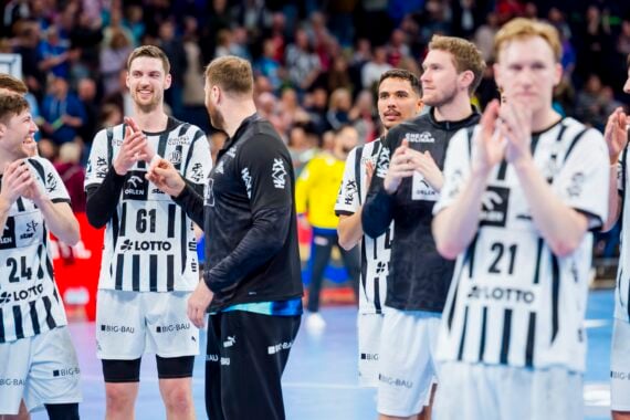 Eine Gruppe männlicher Handballspieler in schwarz-weißen Trikots steht auf einem Spielfeld, klatscht und lächelt, während im Hintergrund eine Menschenmenge zu sehen ist. Einige Spieler sind einander zugewandt und scheinen zu feiern oder etwas anzuerkennen.