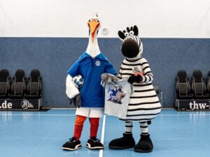 Auf einem blauen Hallenplatz stehen zwei kostümierte Maskottchen: Das eine ist ein Storch in einem blauen Hemd, der einen Ball in der Hand hält, und das andere ist ein Zebra in einem gestreiften Outfit, das ein T-Shirt mit einem verspielten Motiv hochhält.