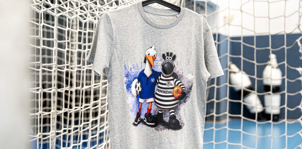 holsteinxthw_titel Ein graues T-Shirt, das an einem schwarzen Kleiderbügel hängt, mit den Comic-Maskottchen eines Storchs in einem blauen Sportoutfit und eines Zebras in einem gestreiften Trikot, das einen Basketball hält, vor dem Hintergrund eines Tornetzes.