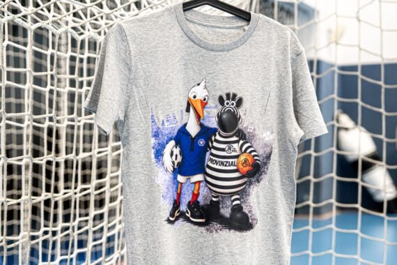Ein graues T-Shirt, das an einem schwarzen Kleiderbügel hängt, mit den Comic-Maskottchen eines Storchs in einem blauen Sportoutfit und eines Zebras in einem gestreiften Trikot, das einen Basketball hält, vor dem Hintergrund eines Tornetzes.
