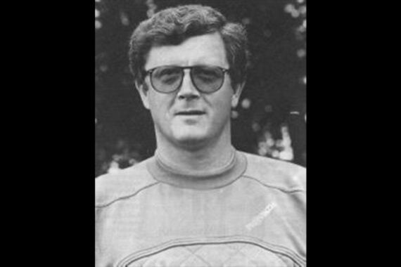 Schwarz-Weiß-Foto eines Mannes, der eine Brille und ein legeres, langärmeliges Hemd trägt. Er hat kurzes, gewelltes Haar und blickt direkt in die Kamera. Im Hintergrund sind Bäume und unscharfes Laub zu sehen.