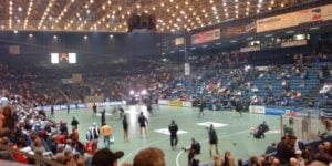 Eine große Sporthalle, die berühmte Ostseehalle, ist mit Zuschauern gefüllt, während sich der THW Kiel unter grellem Flutlicht auf ein Spiel der Handball-Bundesliga vorbereitet. Die Tribünen sind teilweise besetzt, und auf Transparenten wird die reiche Geschichte der Halle gewürdigt.