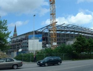 Ein im Bau befindliches Gebäude mit Stahlskelett, umgeben von Gerüsten und einem hohen gelben Kran, überragt Autos und Grünflächen. Im Hintergrund ragt ein Kirchturm in der Nähe der historischen Ostseehalle empor, der Heimat der THW Kiel Handball-Bundesliga-Geschichte.
