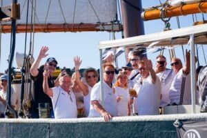 Eine Gruppe von Menschen in einheitlichen weißen Hemden steht auf einem Boot, lächelt und winkt in die Kamera. Einige halten Getränke in der Hand, und die Sonne scheint. Im Hintergrund sind Segel und Takelage zu sehen.