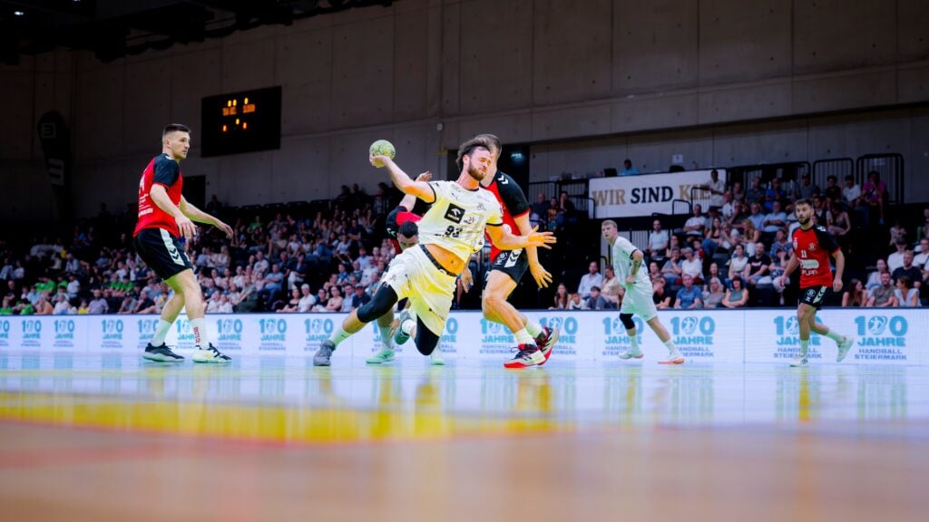 26_Vb_Graz_VorTestspiel Ein Handballspieler in gelbem Trikot stürzt mit dem Ball in der Hand, während Verteidiger in roten und schwarzen Trikots ihn blockieren. Eine Menschenmenge verfolgt das Geschehen von den Tribünen einer hell beleuchteten Halle aus.