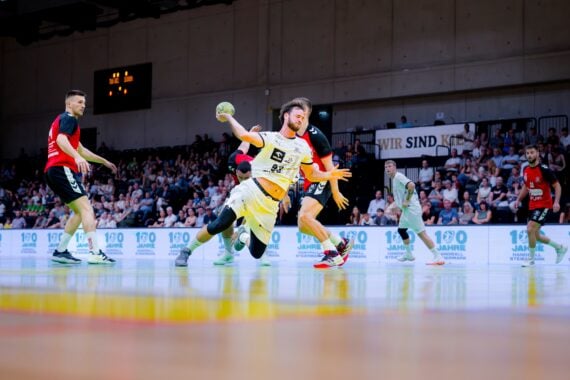 Ein Handballspieler in gelbem Trikot stürzt mit dem Ball in der Hand, während Verteidiger in roten und schwarzen Trikots ihn blockieren. Eine Menschenmenge verfolgt das Geschehen von den Tribünen einer hell beleuchteten Halle aus.