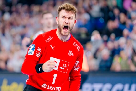 Ein männlicher Handballspieler in einem roten Trikot jubelt begeistert, ballt die Faust und schreit, während im Hintergrund eine verschwommene Menschenmenge zu sehen ist.