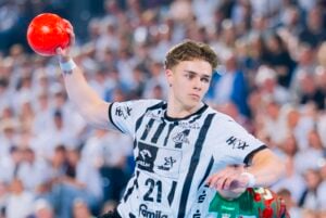 Ein männlicher Handballspieler in einem schwarz-weißen Trikot macht sich während eines Spiels bereit, einen roten Ball zu werfen, im Hintergrund ist eine Zuschauermenge zu sehen.