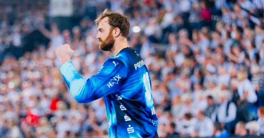Ein Handballspieler in einem blauen Trikot reckt jubelnd die Faust in die Höhe, im Hintergrund ist eine verschwommene Menschenmenge zu sehen.