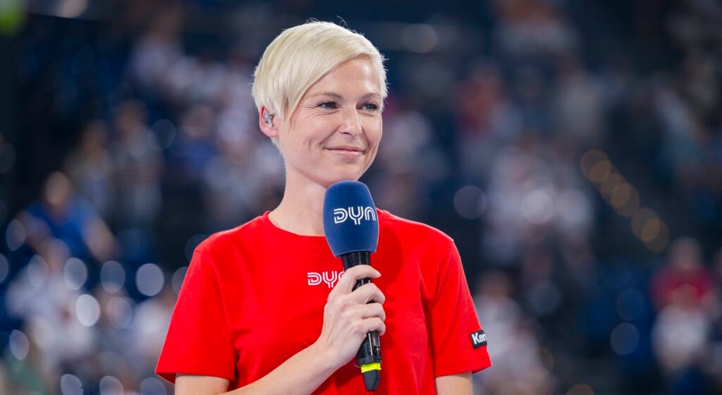 HANDBALL 2023/24: THW Kiel - MT Melsungen Eine lächelnde Person mit kurzen blonden Haaren hält ein Mikrofon in der Hand, trägt ein rotes T-Shirt und steht vor einer verschwommenen Menschenmenge.