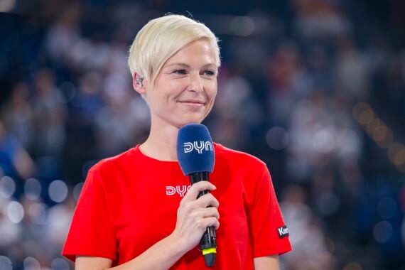 Eine lächelnde Person mit kurzen blonden Haaren hält ein Mikrofon in der Hand, trägt ein rotes T-Shirt und steht vor einer verschwommenen Menschenmenge.
