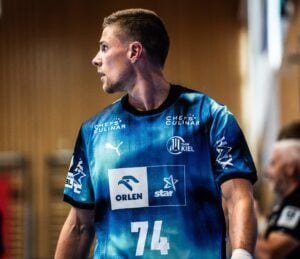 Ein Handballspieler in einem blauen Trikot mit der Nummer 74 schaut während eines Spiels zur Seite. Auf dem Trikot sind Sponsorenlogos zu sehen. Der Hintergrund ist unscharf.