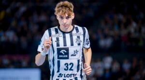 Ein männlicher Handballspieler in einem schwarz-weiß gestreiften Trikot mit der Nummer 32 steht konzentriert auf dem Spielfeld, im Hintergrund ist eine verschwommene Zuschauermenge zu sehen.