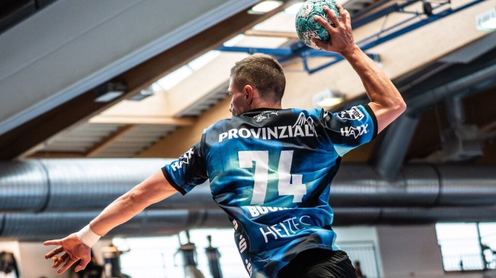 27_PM_Buechner_Titel1 Ein Handballspieler im blauen Trikot mit der Nummer 74 macht sich bereit, den Ball in einer Sporthalle zu werfen, während Sonnenlicht durch die Fenster über ihm hereinströmt.