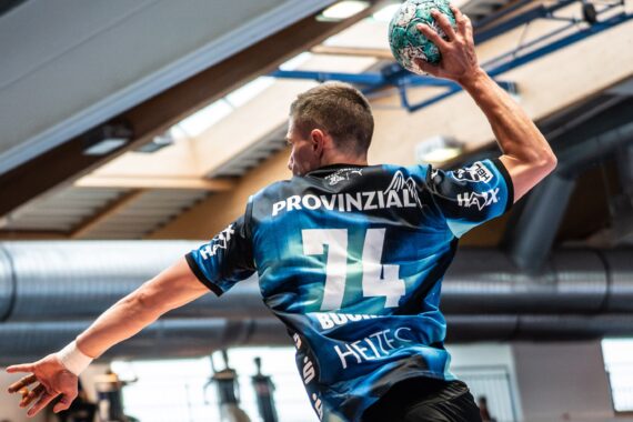 Ein Handballspieler im blauen Trikot mit der Nummer 74 macht sich bereit, den Ball in einer Sporthalle zu werfen, während Sonnenlicht durch die Fenster über ihm hereinströmt.
