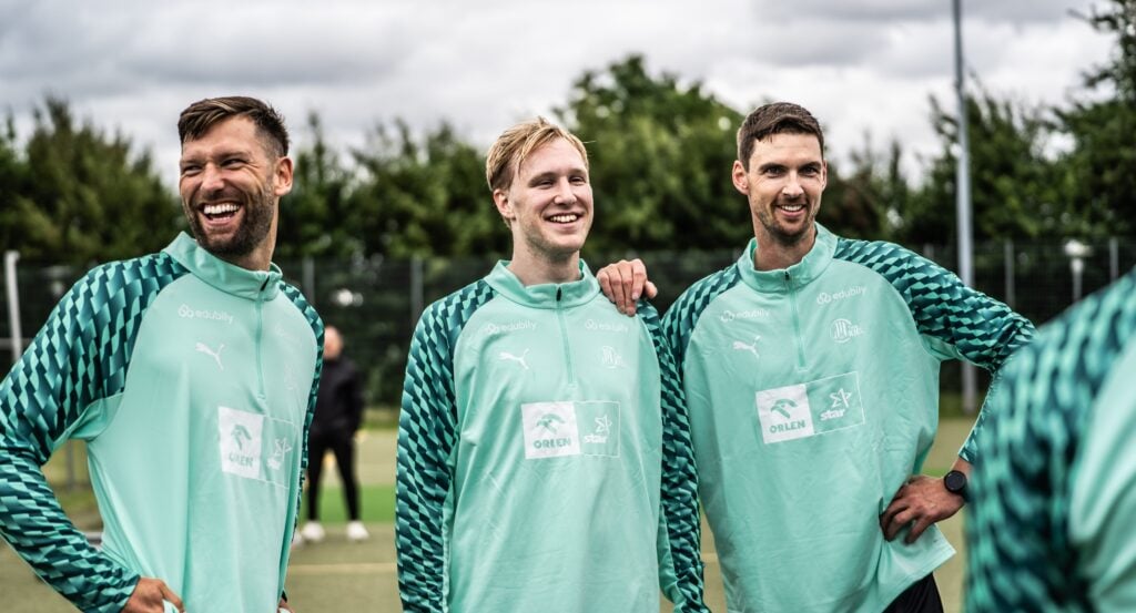 27_Trainingsauftakt-12 Drei Männer in einheitlichen blaugrünen Sportjacken stehen draußen auf einem Feld, lächeln und lachen gemeinsam, im Hintergrund sind Bäume und ein bewölkter Himmel zu sehen.