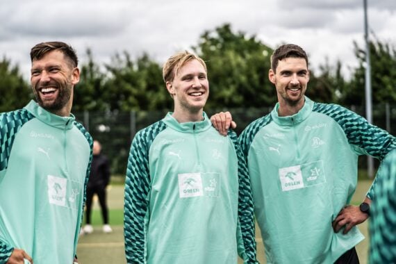 Drei Männer in einheitlichen blaugrünen Sportjacken stehen draußen auf einem Feld, lächeln und lachen gemeinsam, im Hintergrund sind Bäume und ein bewölkter Himmel zu sehen.