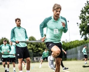 Ein Mann in einem grünen Sportoutfit läuft auf einem Feld, während ein anderer Mann ein Widerstandsband um seine Taille hält. Im Hintergrund stehen weitere Mannschaftskameraden, die alle ähnliche Outfits tragen. Hinter ihnen sind Bäume und ein Zaun zu sehen.