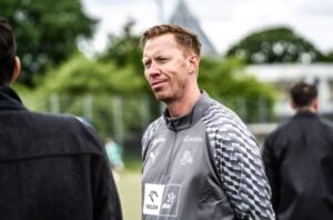 Ein Mann mit kurzen hellbraunen Haaren, der eine graue Sportjacke trägt, steht draußen auf einem Feld und unterhält sich mit jemandem. Im Hintergrund sind Bäume und Menschen verschwommen zu sehen.