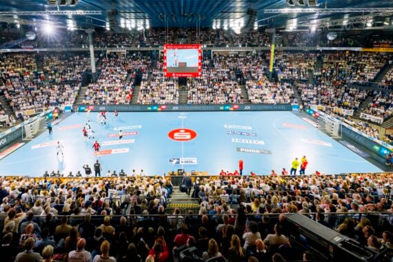Eine große Sporthalle, vollgepackt mit Zuschauern, die ein Handballspiel verfolgen; die Mannschaften spielen auf einem blauen Spielfeld, und über dem Spielfeld befindet sich eine Anzeigetafel.