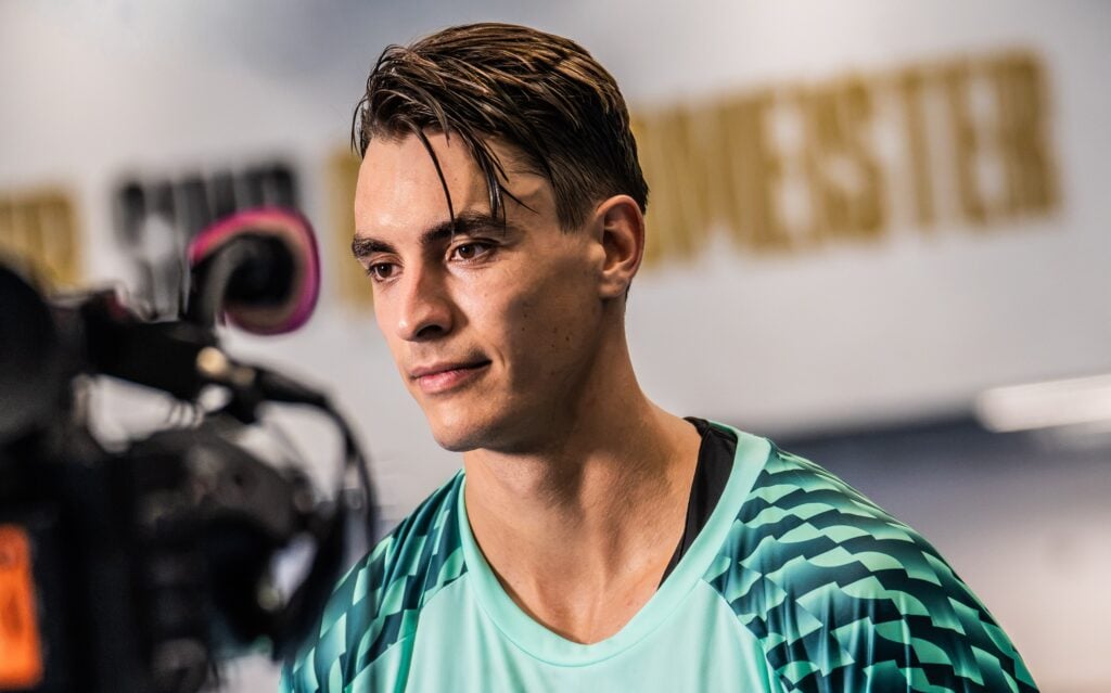 27_koeter_Kapitän_Titel Ein junger Mann, der ein gemustertes, blaugrünes Sportshirt trägt, blickt nach vorne, während er von einer Kamera in einem Innenraum mit unscharfem Hintergrundtext gefilmt wird.