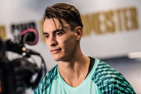 Ein junger Mann, der ein gemustertes, blaugrünes Sportshirt trägt, blickt nach vorne, während er von einer Kamera in einem Innenraum mit unscharfem Hintergrundtext gefilmt wird.