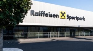 Die Außenansicht des Raiffeisen-Sportparks, eines modernen Gebäudes mit großen Glasfenstern und einem Schild mit dem Raiffeisen-Logo an der Fassade. Auf der linken Seite des Bildes ist ein Baum zu sehen.