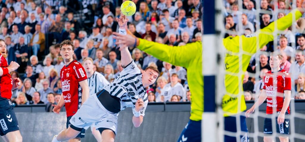 Ein Handballspieler in einem weiß gestreiften Trikot wirft den Ball in Richtung Tor, während sich der Torwart in Gelb darauf vorbereitet, den Ball abzuwehren; die Spieler und eine große Zuschauermenge verfolgen das Geschehen gespannt.