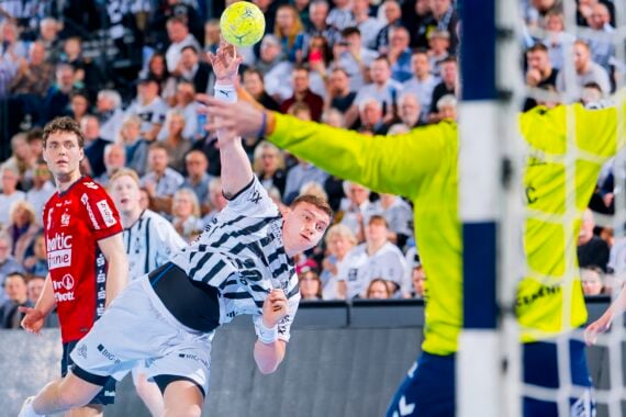 Ein Handballspieler in einem weiß gestreiften Trikot wirft den Ball in Richtung Tor, während sich der Torwart in Gelb darauf vorbereitet, den Ball abzuwehren; die Spieler und eine große Zuschauermenge verfolgen das Geschehen gespannt.