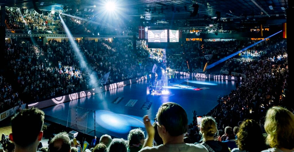 Eine große Sporthalle, vollgepackt mit Zuschauern, die auf eine hell erleuchtete Bühne auf einem Sportplatz blicken, wobei Scheinwerfer die Moderatoren in der Mitte anstrahlen.