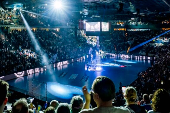 Eine große Sporthalle, vollgepackt mit Zuschauern, die auf eine hell erleuchtete Bühne auf einem Sportplatz blicken, wobei Scheinwerfer die Moderatoren in der Mitte anstrahlen.