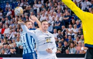 Ein Handballspieler im weißen Trikot ist gerade dabei, den Ball zu werfen, während ein blau gestreifter Verteidiger und ein gelb gekleideter Torwart reagieren. Im Hintergrund verfolgt eine Menschenmenge das Geschehen gespannt.