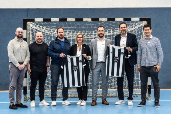 Sieben Personen stehen vor einem Hallenfußballtor, zwei von ihnen halten schwarz-weiß gestreifte Sporttrikots in den Händen. Sie tragen Business-Casual-Kleidung und lächeln für das Foto.
