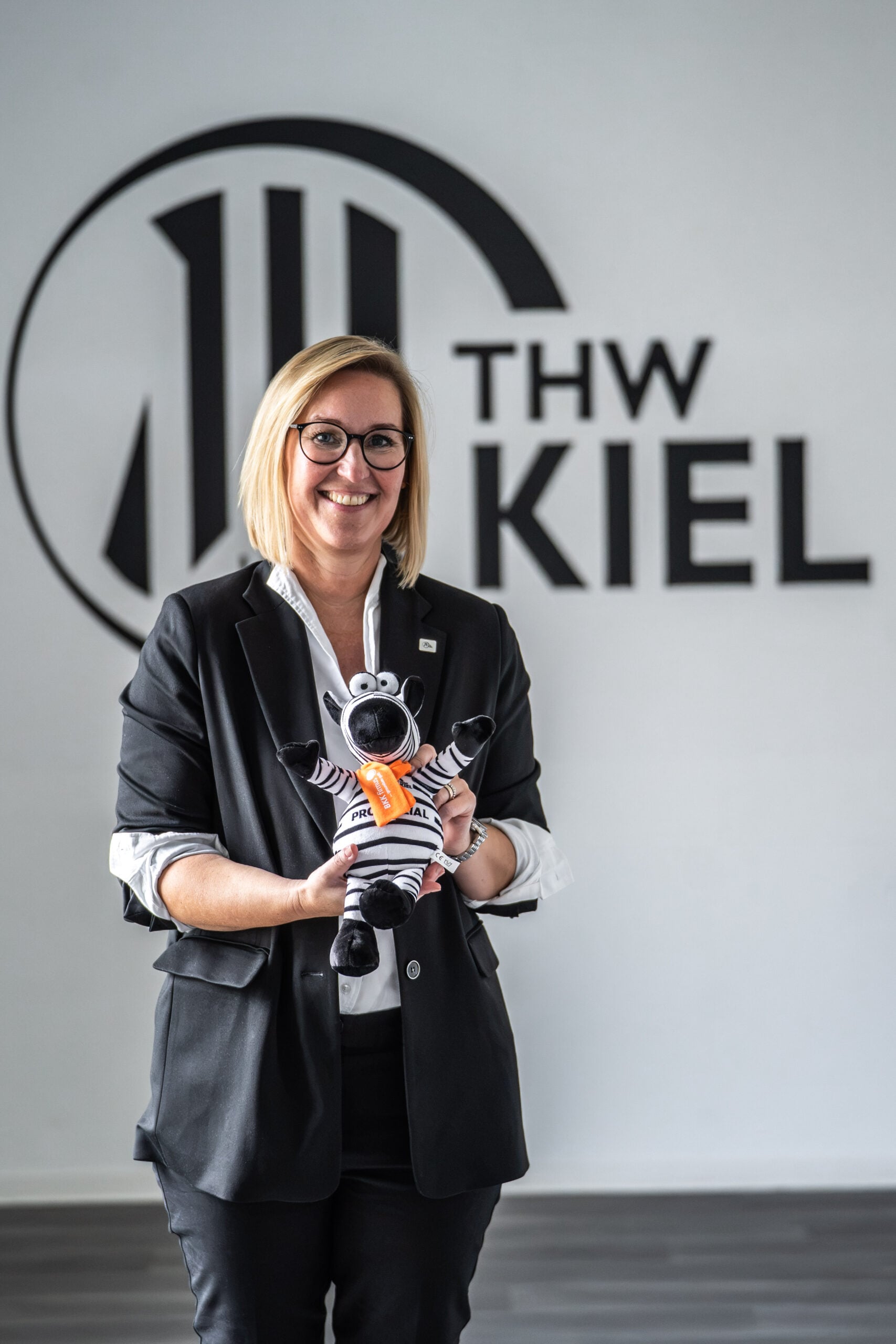 Eine lächelnde Person in einem schwarzen Anzug und mit Brille steht in einem Raum und hält ein Zebra-Plüschtier in der Hand. Hinter ihr ist eine Wand mit dem Logo und dem Schriftzug „THW KIEL.“ zu sehen.