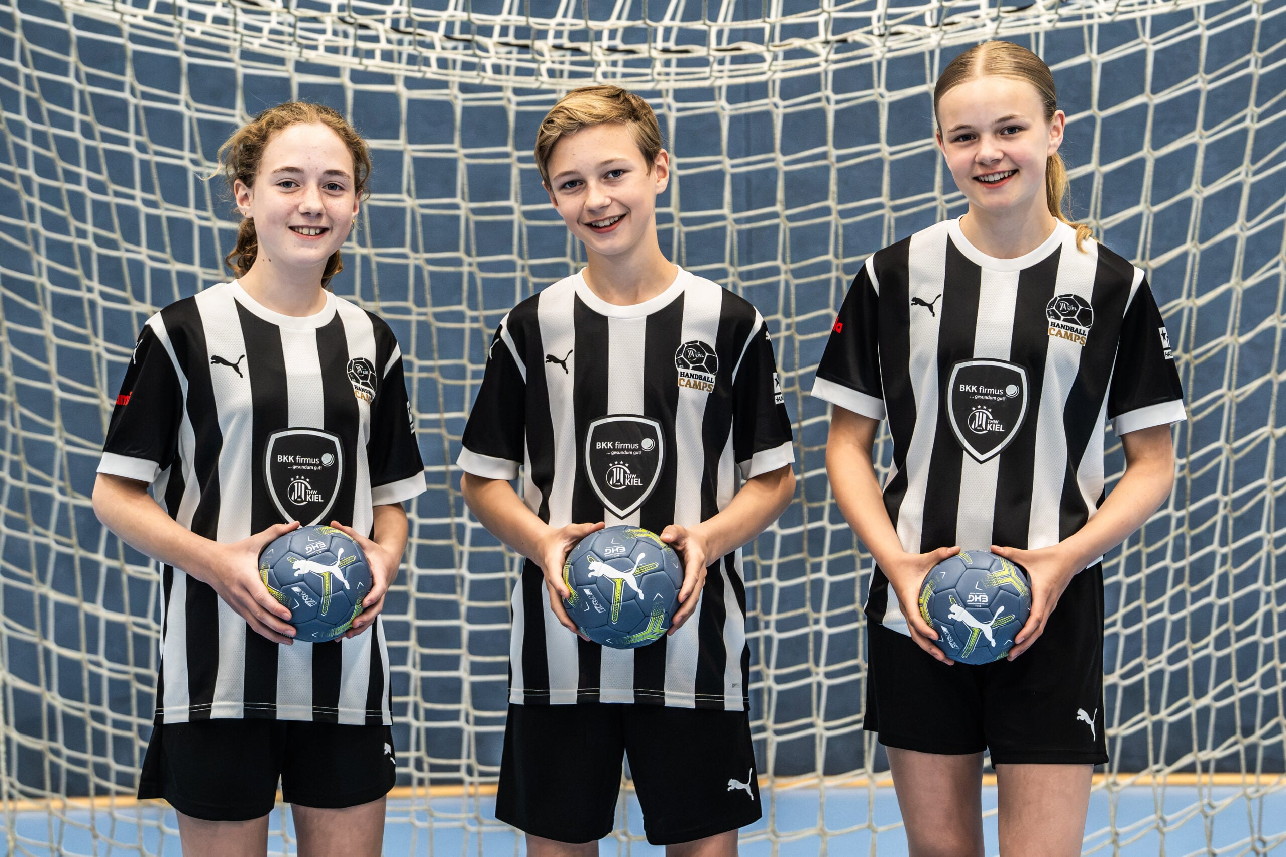 Drei junge Handballspieler in schwarz-weiß gestreiften Trikots stehen vor einem Tor, halten jeweils einen blauen Handball in der Hand und lächeln in die Kamera.