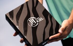 Eine Person hält eine schwarze Schachtel mit zebraartigen Streifen und dem THW-Kiel-Logo in Weiß auf dem Deckel in der Hand. Zu sehen sind nur die Hände der Person und ein Teil ihres Arms.
