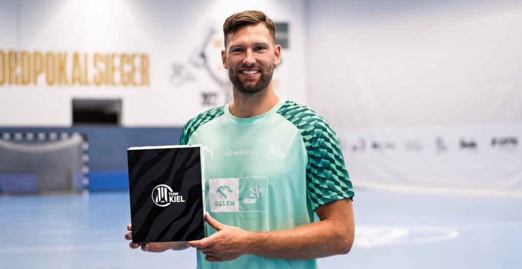 Box_Harald-4 Ein Mann in einem grünen Sportshirt hält eine schwarz-weiße THW-Kiel-Kiste in der Hand und steht auf einer Sporthalle mit blauem Bodenbelag und unscharfen Bannern im Hintergrund.