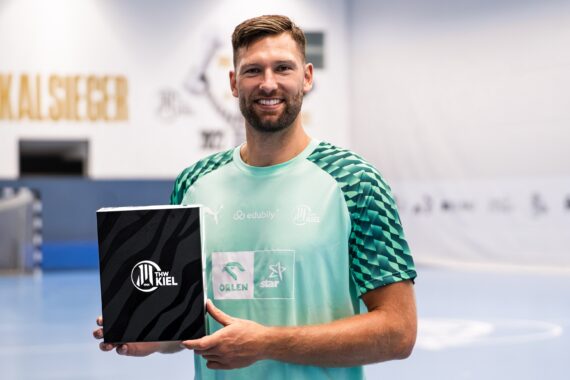 Ein Mann in einem grünen Sportshirt hält eine schwarz-weiße THW-Kiel-Kiste in der Hand und steht auf einer Sporthalle mit blauem Bodenbelag und unscharfen Bannern im Hintergrund.