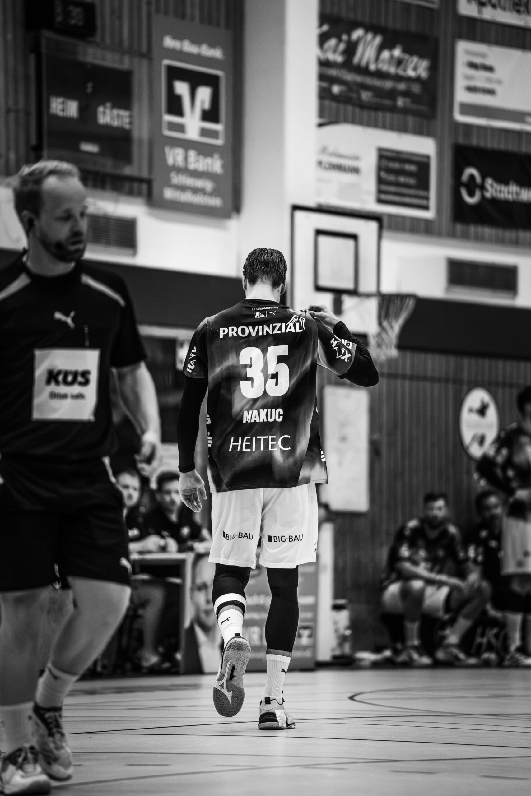 Ein Handballspieler mit der Rückennummer 35 läuft auf einer Halle, umgeben von seinen Mannschaftskollegen und Werbeplakaten an der Wand. Das Schwarz-Weiß-Foto hält den Moment fest, in dem der neue Trainer Börge Lund beim THW Kiel vorgestellt wurde.
