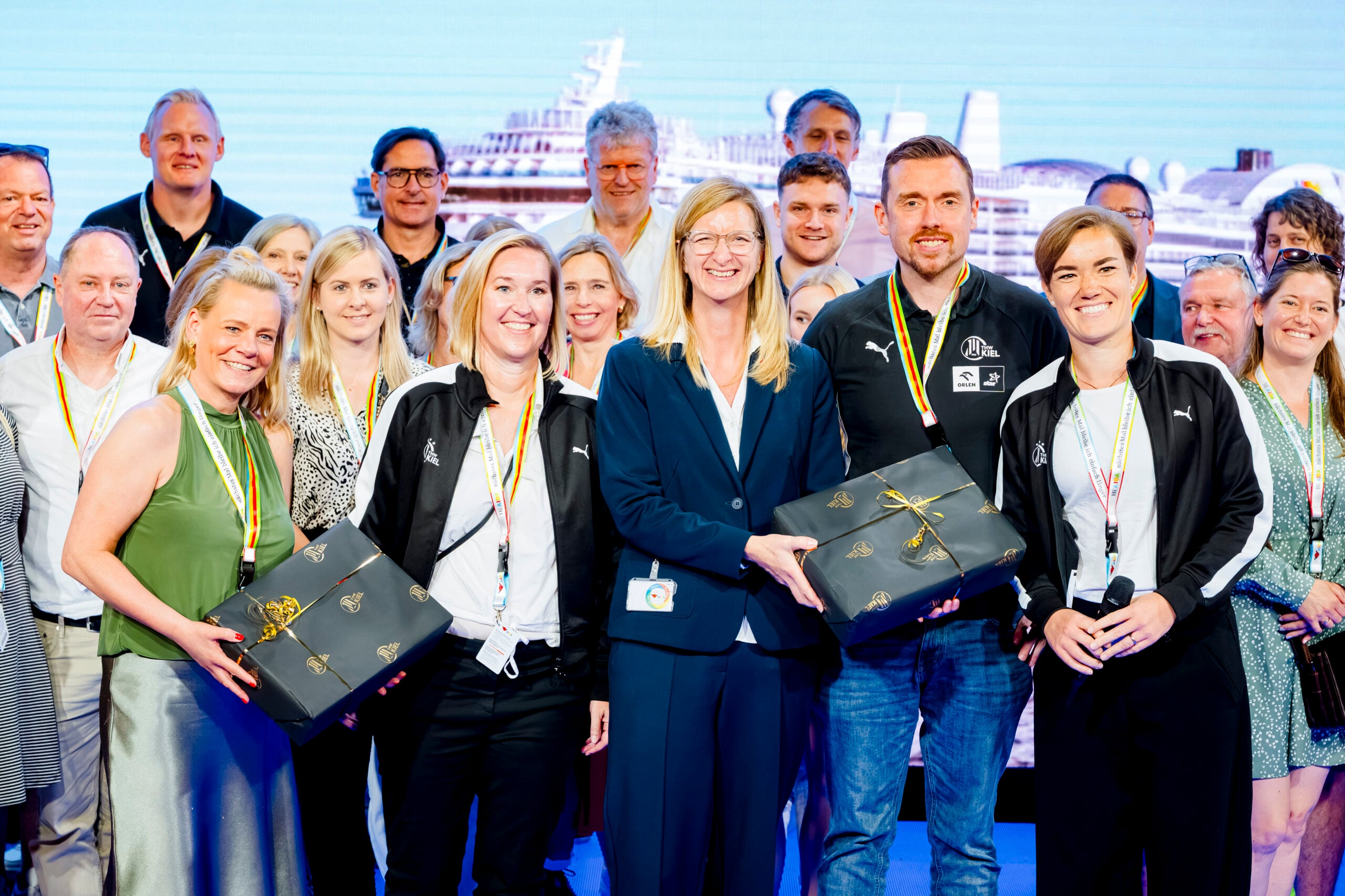 Eine große Gruppe lächelnder Menschen posiert gemeinsam auf der Bühne, einige halten schwarze Geschenkboxen in den Händen. Sie tragen Namensschilder und Schlüsselbänder, hinter ihnen ist ein Schiff oder eine Fähre als Kulisse zu sehen. Die feierliche Atmosphäre spiegelt die Vorfreude auf die kommende Handball-Bundesliga-Saison 2026/2027 des THW Kiel und die Vorstellung ihres neuen Trainers, Börge Lund, wider.