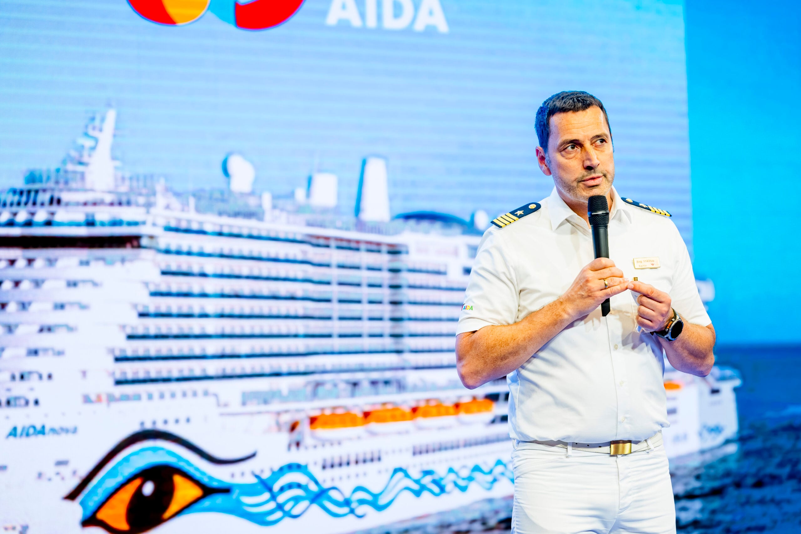 Ein Mann in einer weißen Kapitänsuniform spricht auf der Bühne und hält ein Mikrofon in der Hand. Hinter ihm ist ein großes Bild eines Kreuzfahrtschiffs sowie die Logos von AIDA und Mastercard zu sehen. Im Rahmen der Veranstaltung wird außerdem bekannt gegeben, dass Börge Lund neuer Trainer des THW Kiel in der Handball-Bundesliga-Saison 2026/2027 wird.