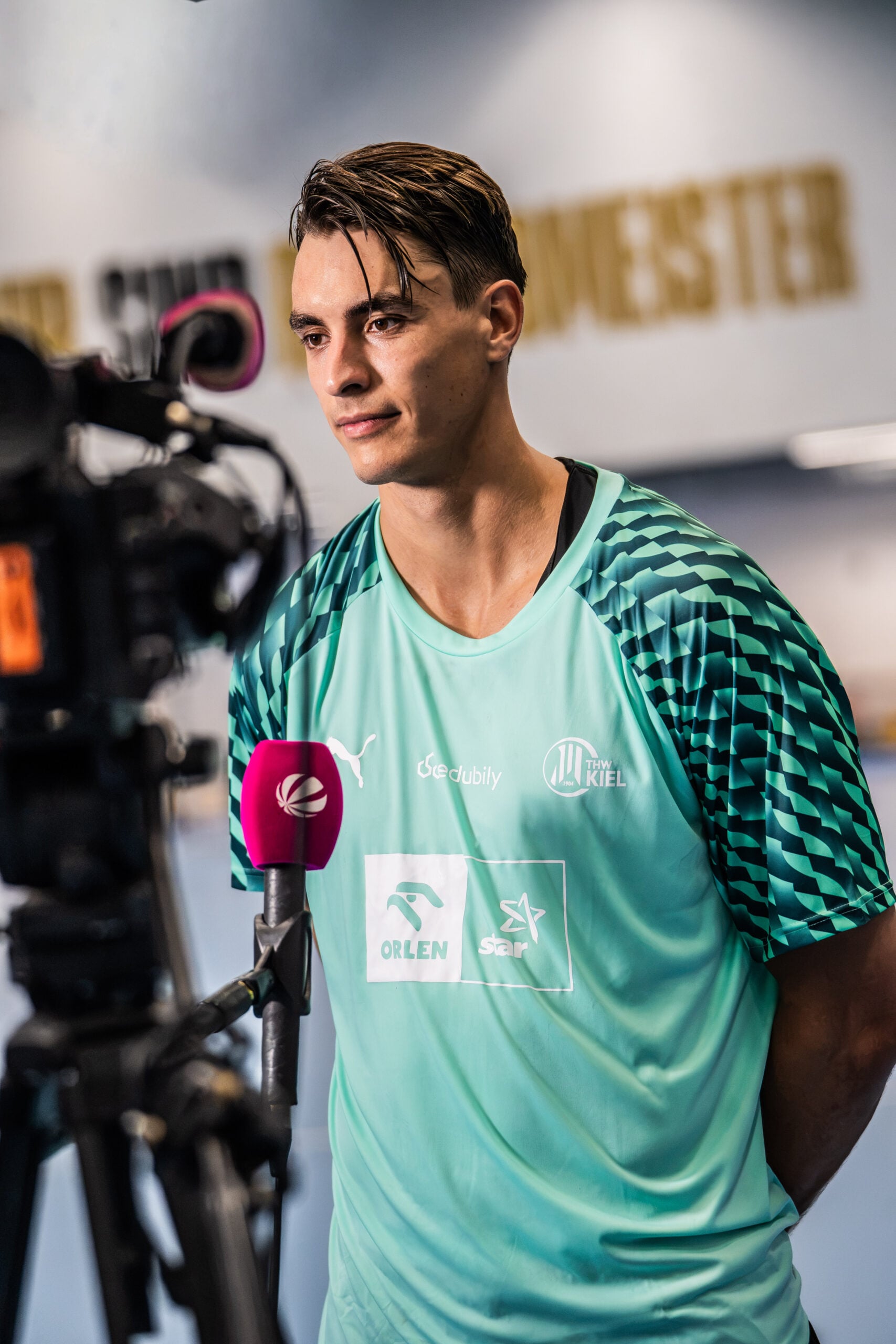 Ein männlicher Sportler in einem blaugrünen Sporttrikot steht vor einer Kamera und einem Mikrofon und gibt in einem Innenraum ein Interview, während im Hintergrund goldfarbene Schriftzüge verschwommen zu sehen sind. Der neue Trainer Börge Lund wurde beim THW Kiel vorgestellt.