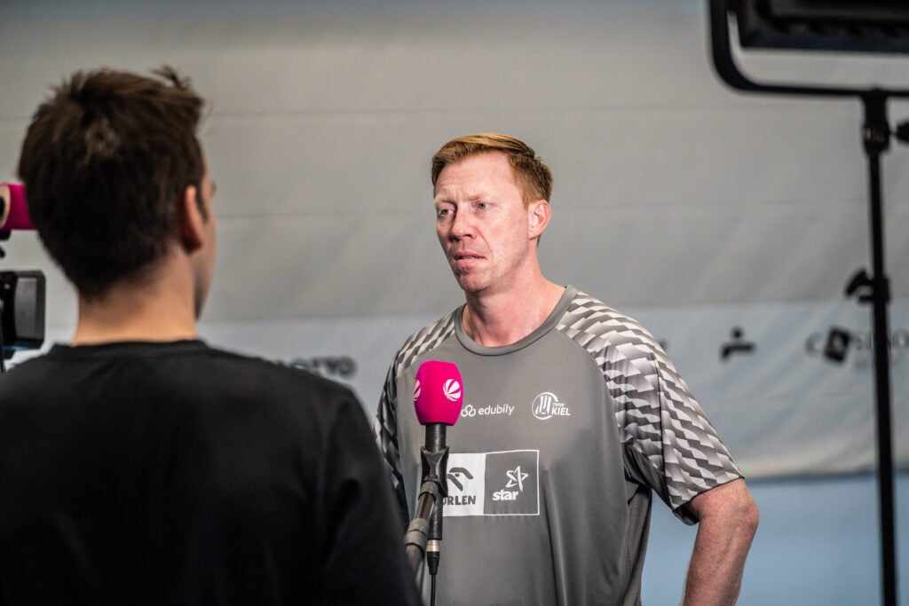 Medien-Training-4 Ein Mann in einem Sporthemd wird mit einem Mikrofon vor sich interviewt und spricht mit einer Person, die der Kamera den Rücken zuwendet. Die Szene spielt in einem Innenraum, als Neu-Trainer Börge Lund beim THW Kiel vorgestellt wurde.