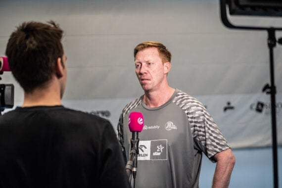 Ein Mann in einem Sporthemd wird mit einem Mikrofon vor sich interviewt und spricht mit einer Person, die der Kamera den Rücken zuwendet. Die Szene spielt in einem Innenraum, als Neu-Trainer Börge Lund beim THW Kiel vorgestellt wurde.