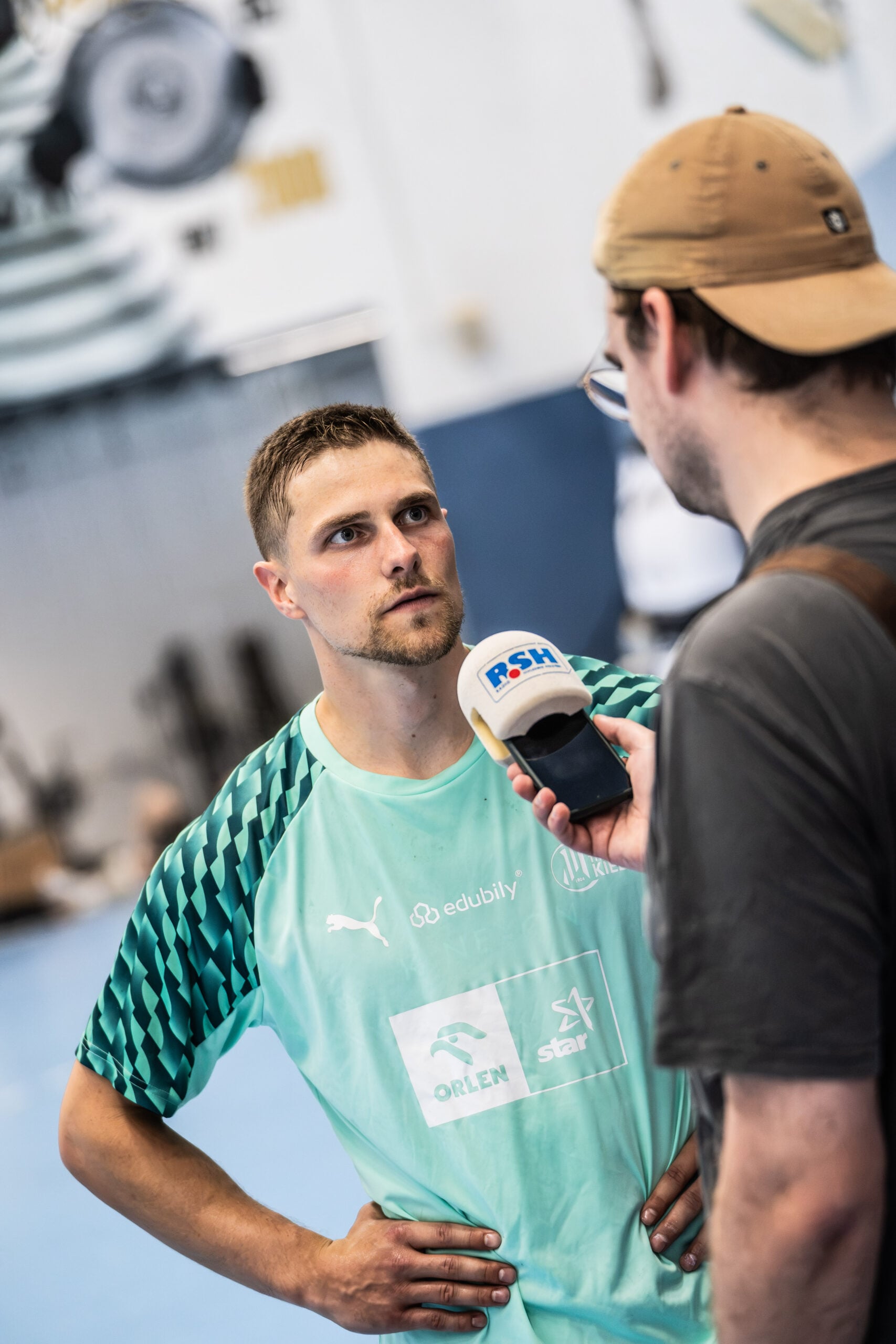 Ein Mann in einem blaugrünen Sporttrikot wird von einem anderen Mann interviewt, der ein Mikrofon mit der Aufschrift „RSH“ in der Hand hält; die Szene spielt in einer Sporthalle, als der neue Trainer Börge Lund beim THW Kiel vorgestellt wurde.