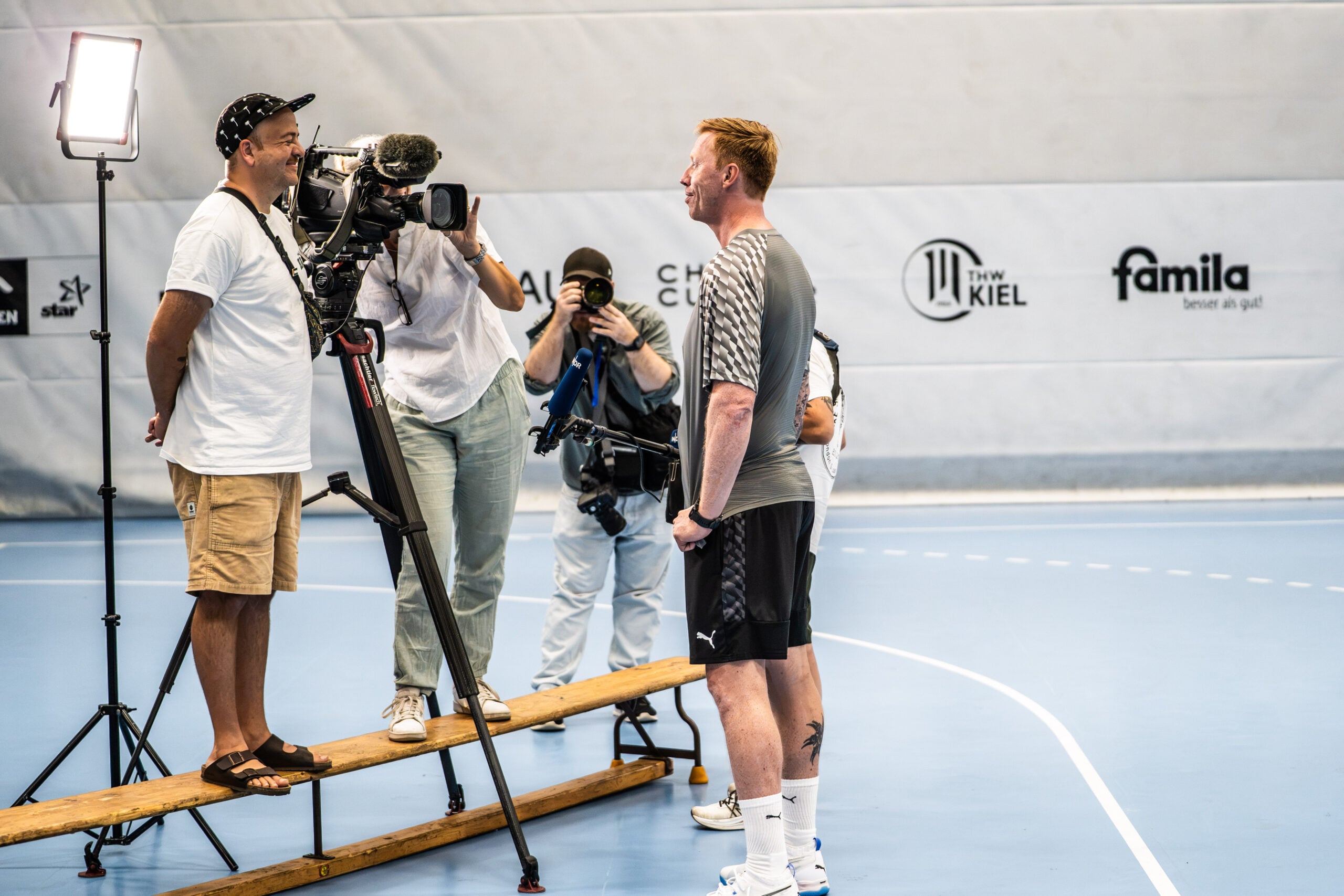 Ein Mann in Sportkleidung steht auf einem blauen Hallenplatz und wird von einem Kamerateam interviewt. Zwei Personen mit Kameras und eine Person mit einem Mikrofon richten ihre Aufmerksamkeit auf ihn. Helle Scheinwerfer beleuchten die Szene, als der neue Trainer Börge Lund beim THW Kiel vorgestellt wurde.