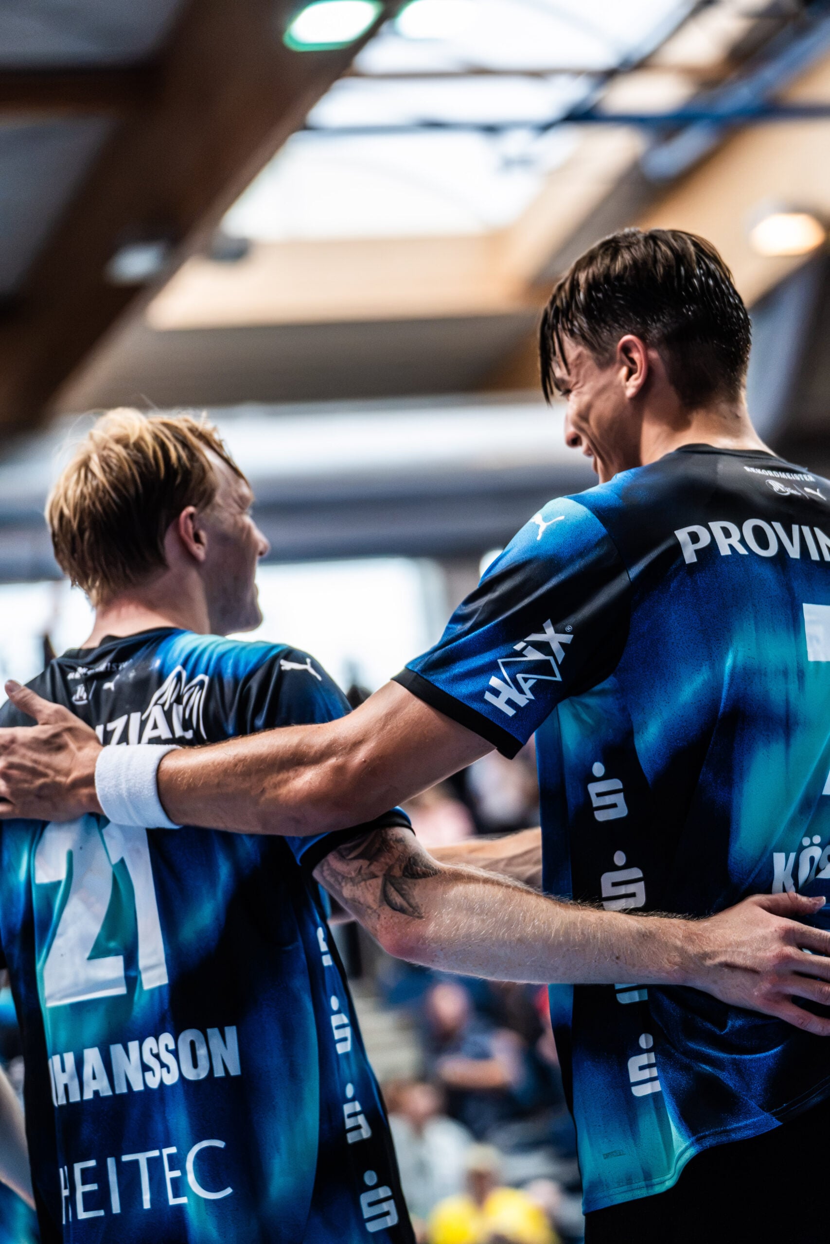 Zwei männliche Sportler in blauen Trikots, die sich umarmen, lächeln und während einer Hallensportveranstaltung feiern. Im Hintergrund sind ein verschwommenes Publikum und helle Scheinwerfer zu sehen, die die Begeisterung bei der Vorstellung des neuen Trainers Börge Lund beim THW Kiel einfangen.