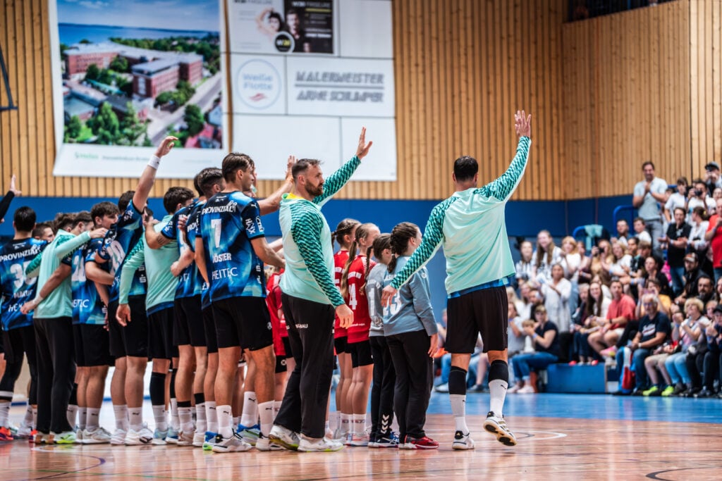 Spiel-35 Eine Sportmannschaft in blau-weißen Trikots winkt einer jubelnden Menge in einer Sporthalle zu, wobei einige Spieler neben Kindern stehen. Im Hintergrund sitzen und stehen die Zuschauer, während der neue Trainer Börge Lund beim THW Kiel vorgestellt wurde.
