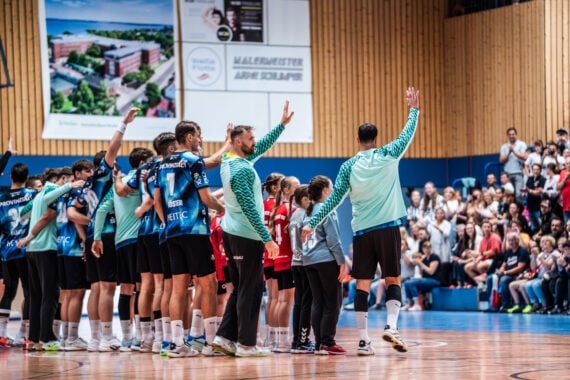 Eine Sportmannschaft in blau-weißen Trikots winkt einer jubelnden Menge in einer Sporthalle zu, wobei einige Spieler neben Kindern stehen. Im Hintergrund sitzen und stehen die Zuschauer, während der neue Trainer Börge Lund beim THW Kiel vorgestellt wurde.